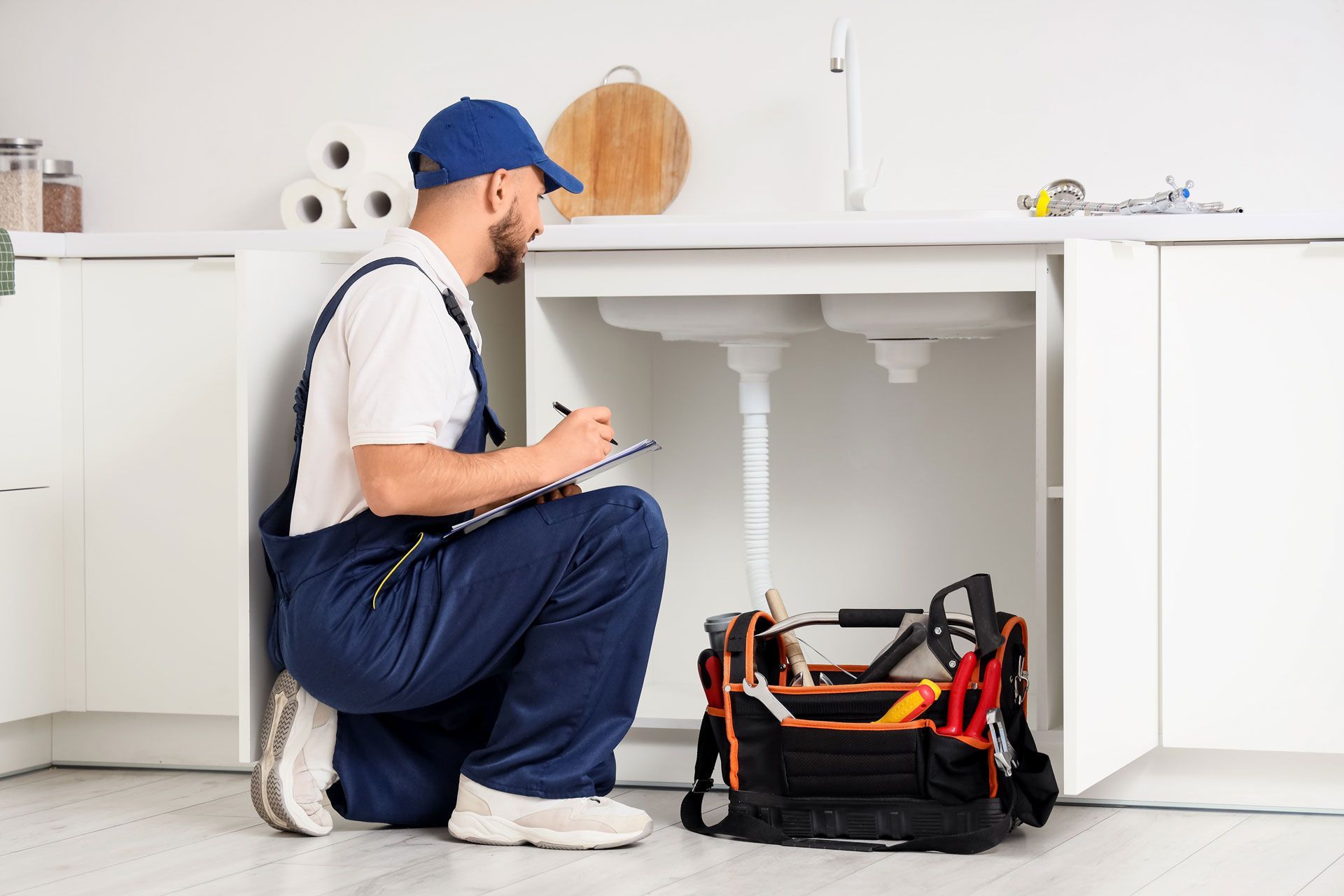 Plumber kneeling under a kitchen sink, looking at pipes and taking notes from a clipboard.