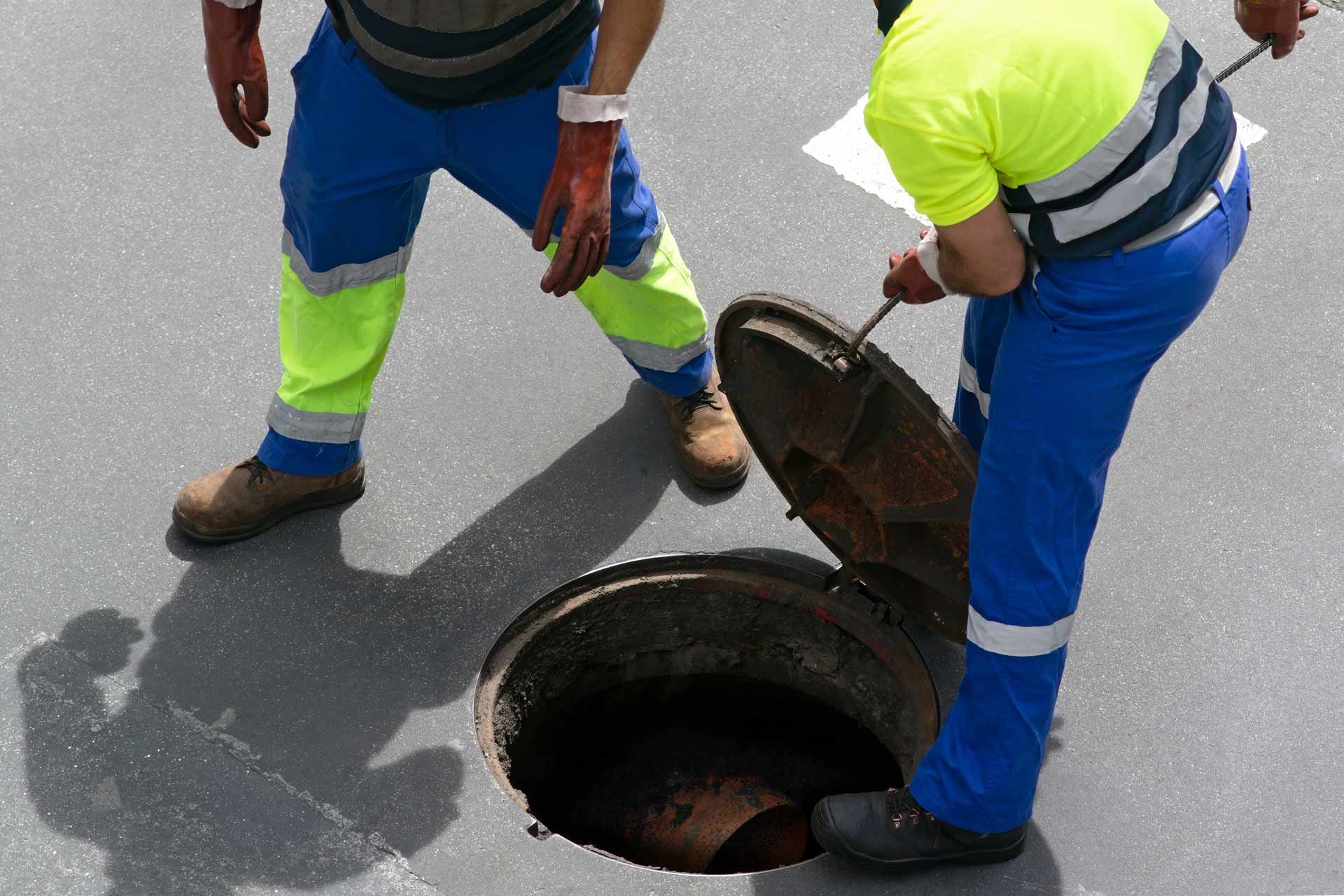 Two workers in safety gear opening a manhole on a paved surface.