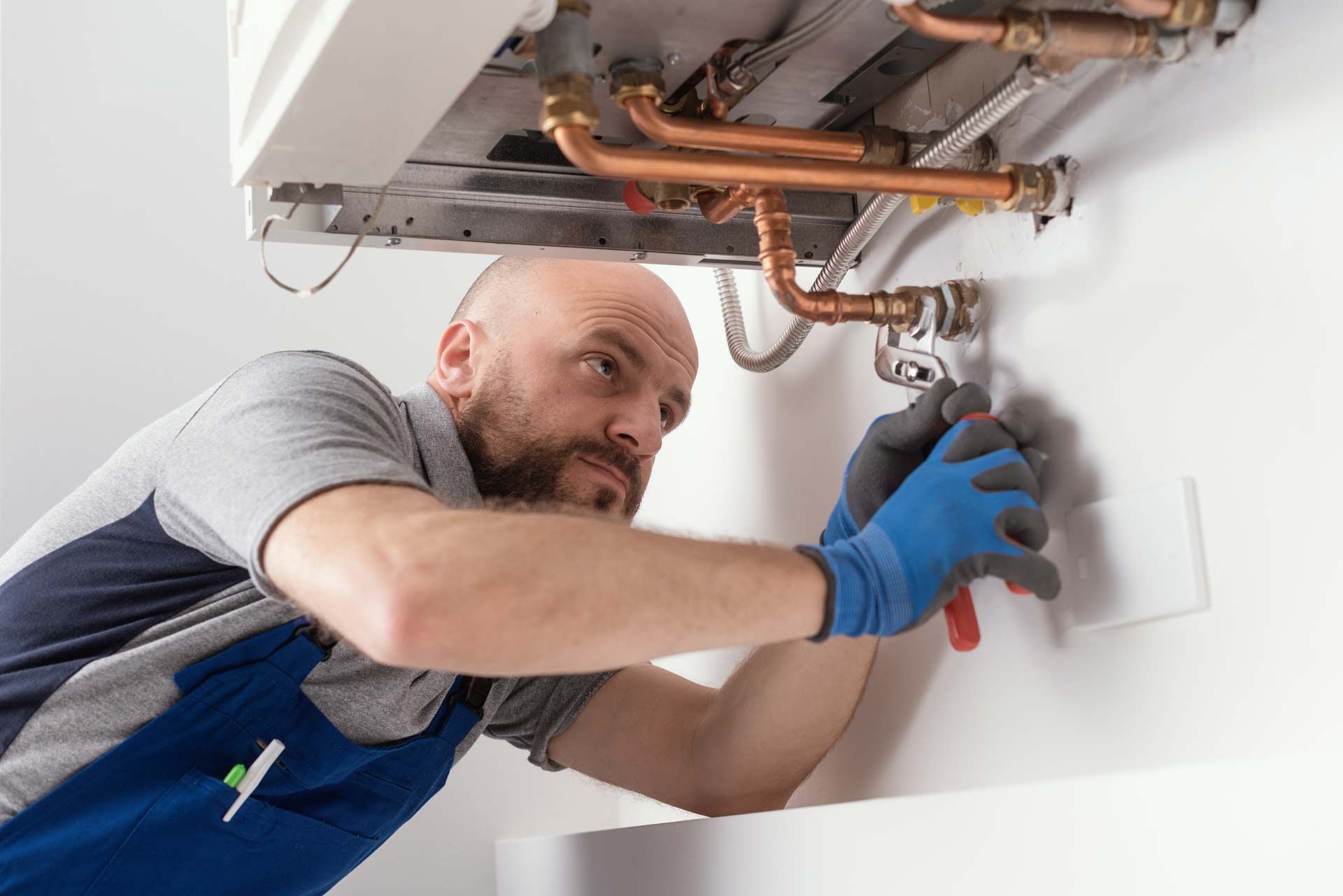 Plumber wearing blue gloves working on a white boiler attached to a white wall, using pliers.