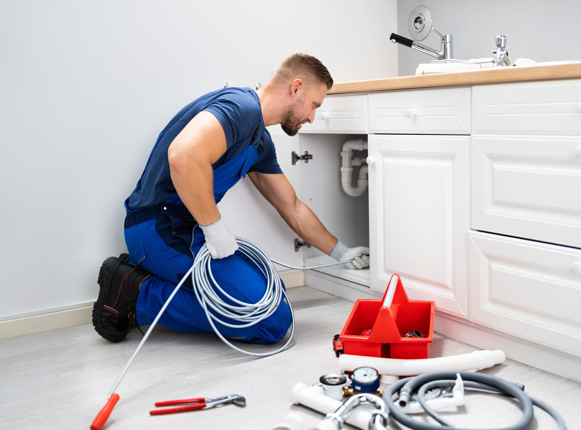Plumber kneels under sink, using a snake tool.