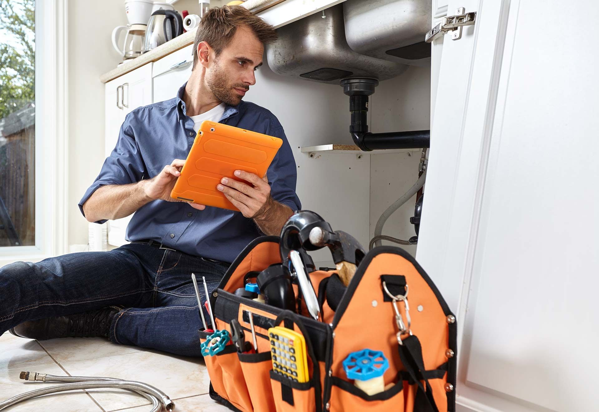 Plumber examines pipes under a kitchen sink, using a tablet, with a toolbox nearby.