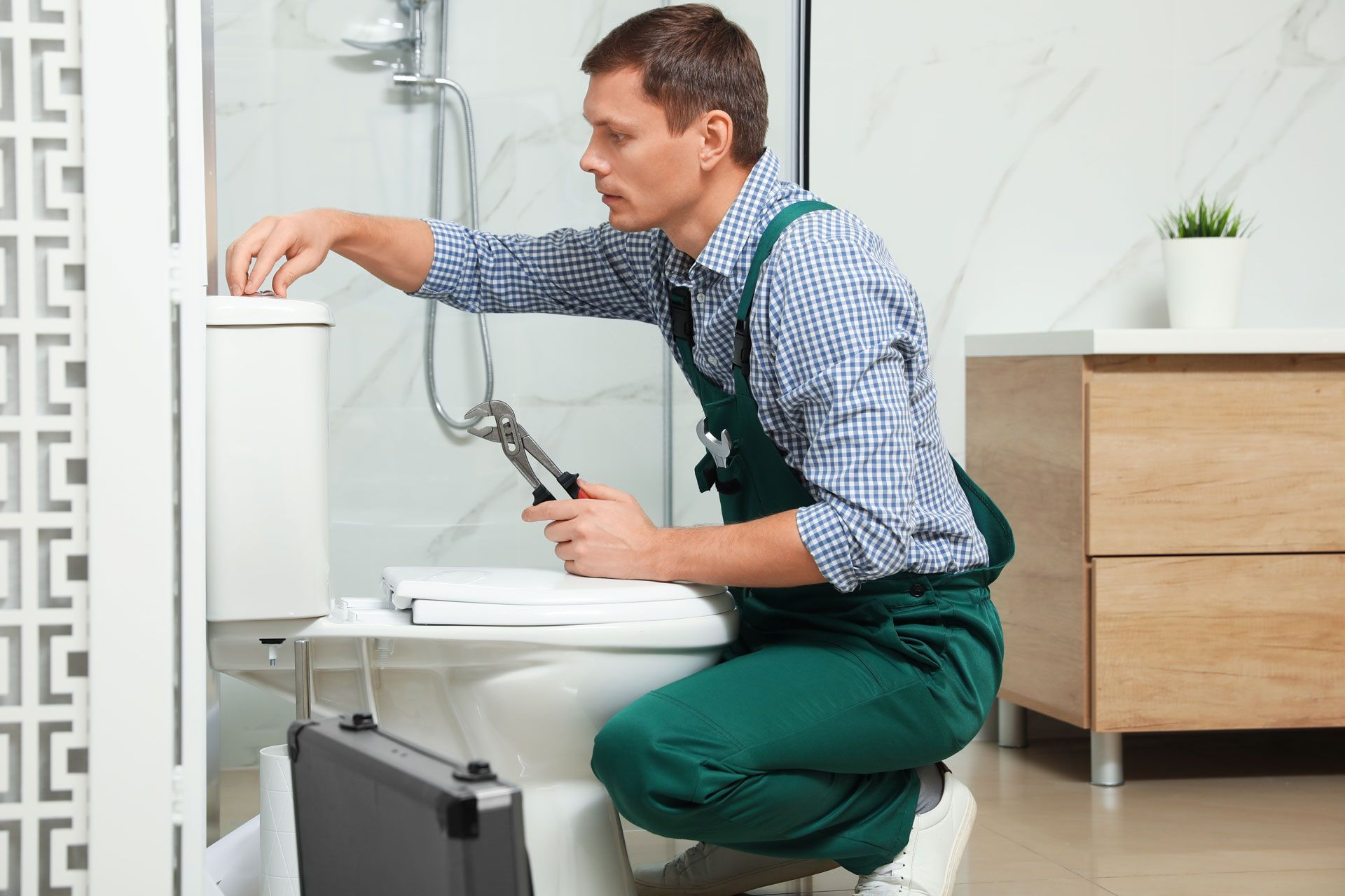 Plumber in green overalls kneels, working on a toilet with a wrench in a bathroom.