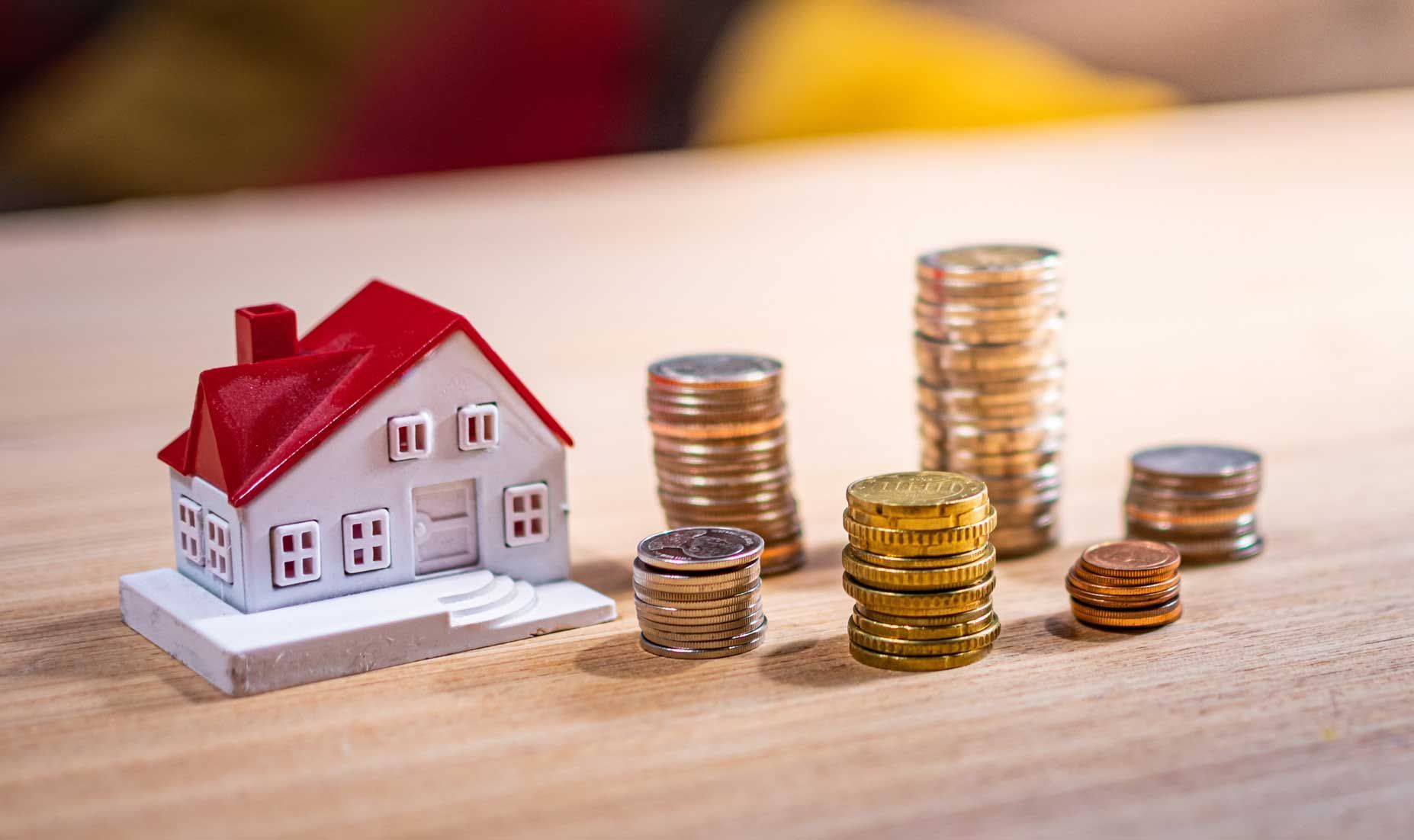 Miniature house next to stacks of coins, on a wooden surface.