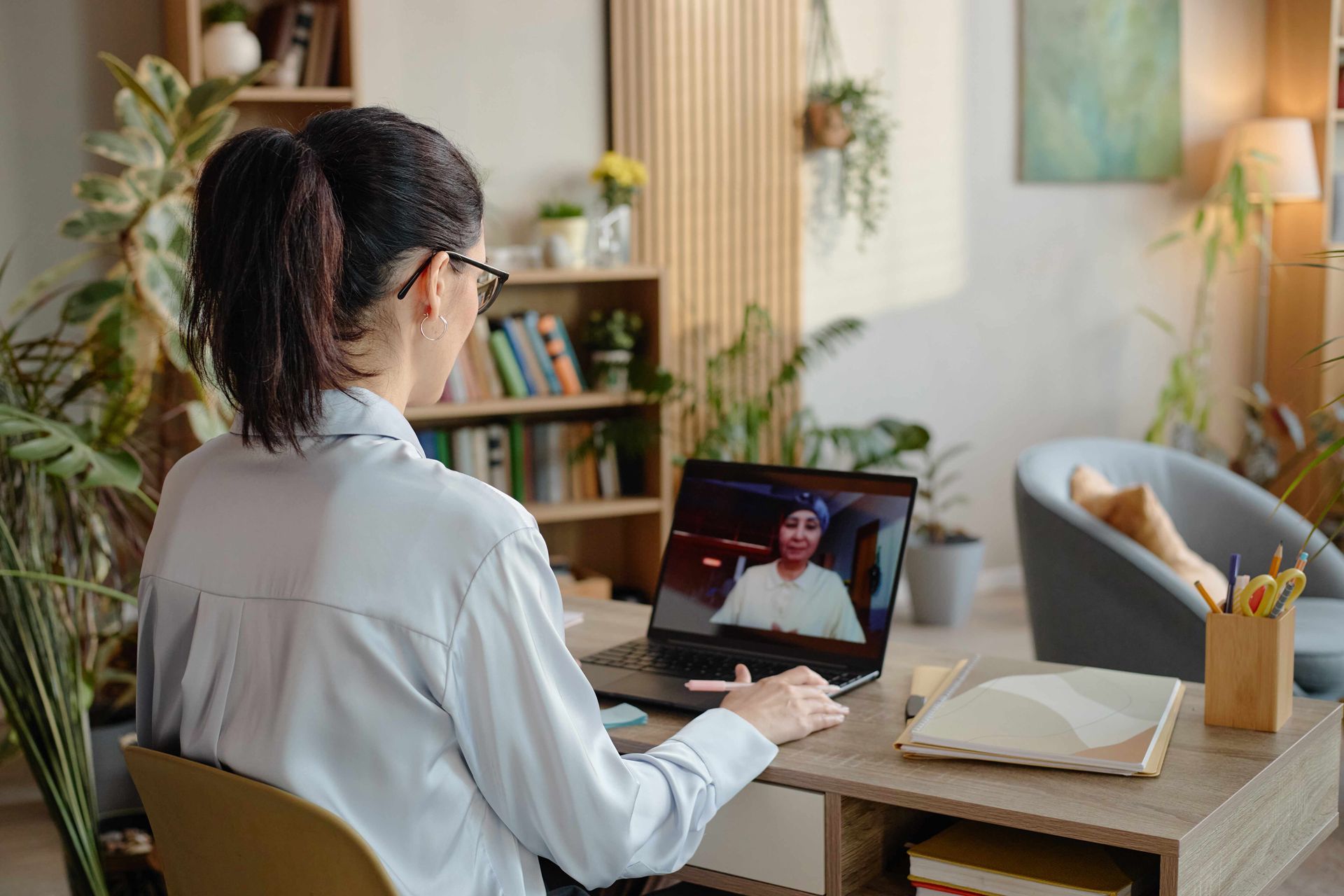 Person working on a laptop at a dining table during a video call, with books and a lamp nearby