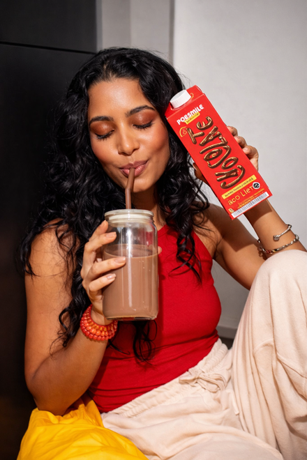 Mulher bebendo leite com chocolate, segurando a caixinha, vestindo blusa vermelha e calça bege.