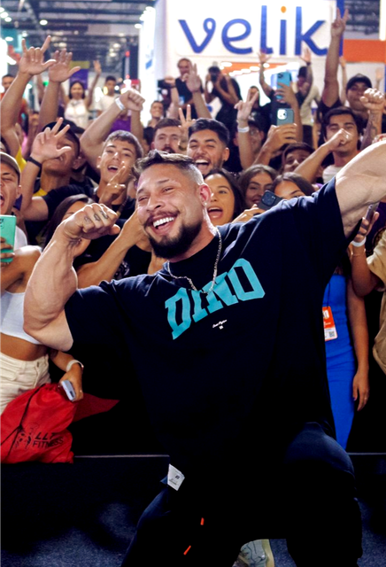 Homem de camisa preta falando ao microfone, segurando um controle remoto, com um cenário de palco ao fundo.