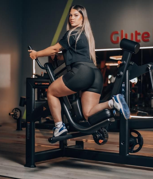 Mulher exercitando os glúteos em um aparelho na academia. Ela está usando uma blusa e shorts pretos, além de tênis azuis e brancos.