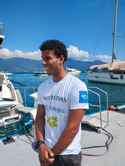 Homem de camisa preta falando ao microfone, segurando um controle remoto, com um cenário de palco ao fundo.