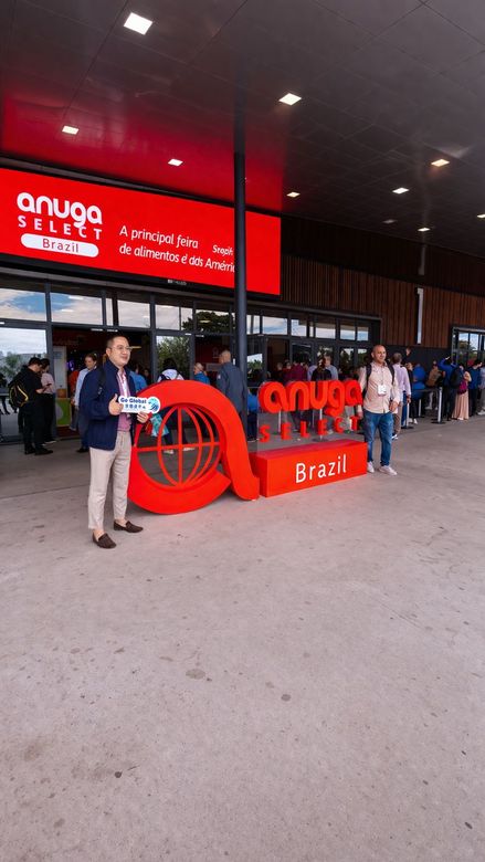 Homem de camisa preta falando ao microfone, segurando um controle remoto, com um cenário de palco ao fundo.