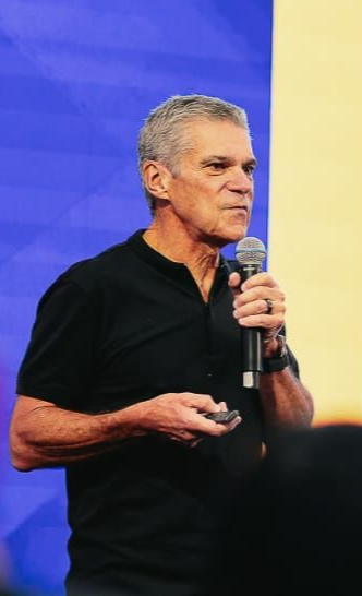 Homem de camisa preta falando ao microfone, segurando um controle remoto, com um cenário de palco ao fundo.