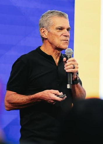 Homem segurando um microfone, falando para uma plateia. Ele está vestindo uma camisa preta.