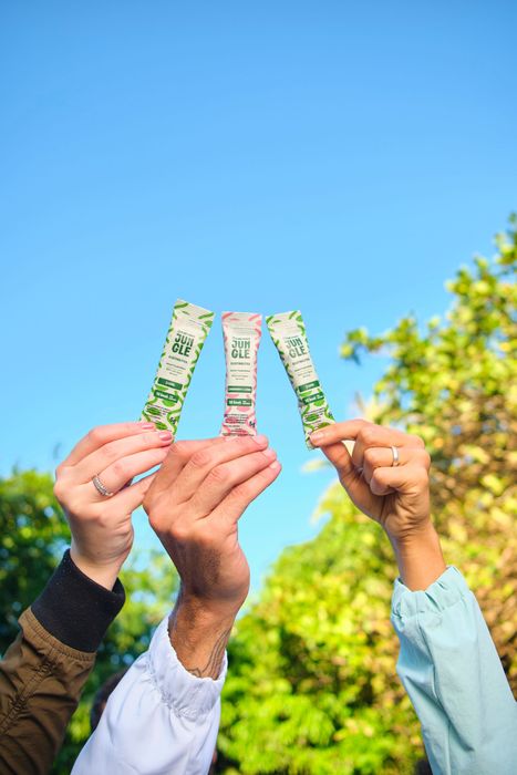 Três mãos segurando pacotes contra um céu azul, cercadas por folhagem verde.
