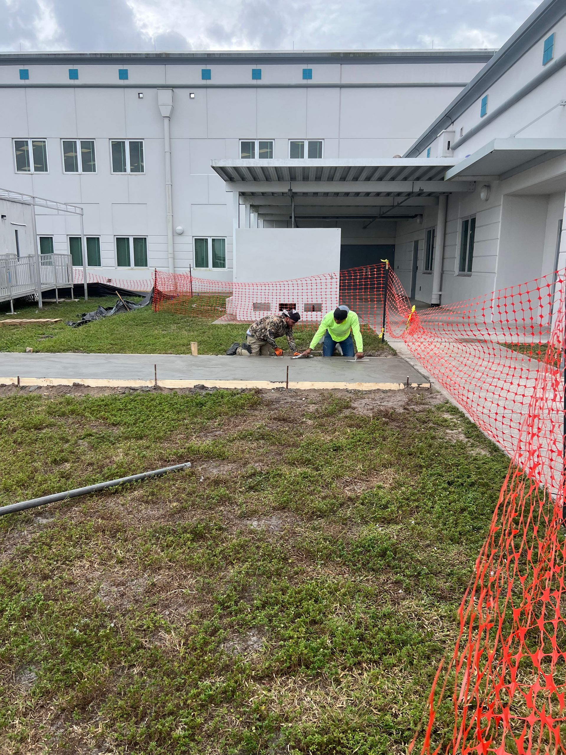 Construction worker kneeling on concrete, near building with orange safety fencing, laying concrete walkway.