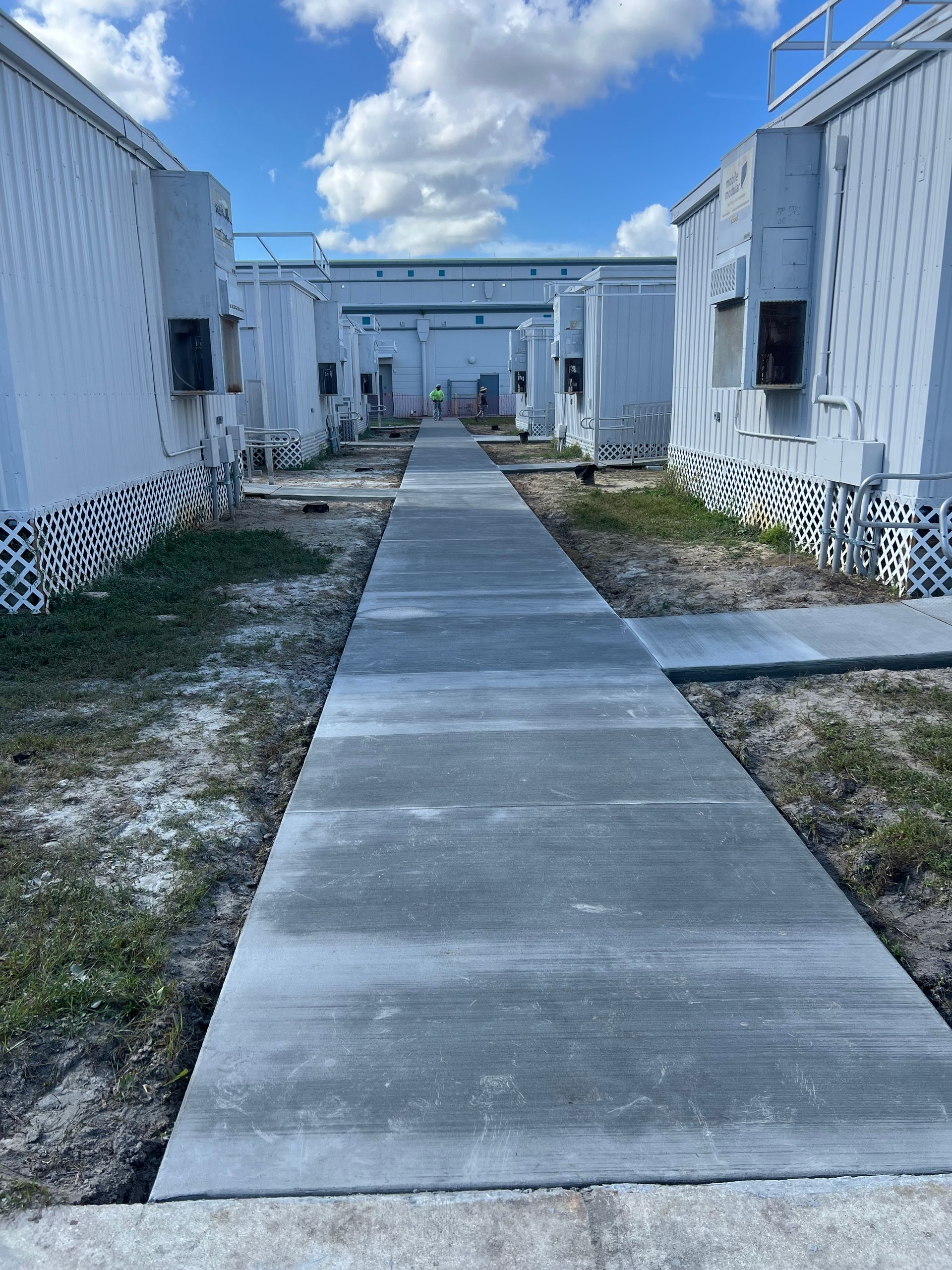 Concrete walkway between rows of white, modular buildings under a blue sky.