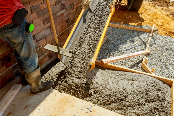 Pouring concrete for a sidewalk; worker uses tools, wooden frame, brick wall in background.