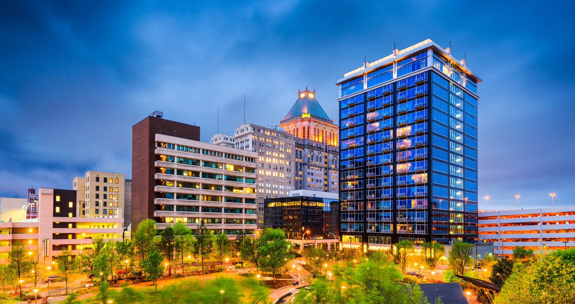 City skyline at dusk, featuring tall buildings with illuminated windows and a park in the foreground.