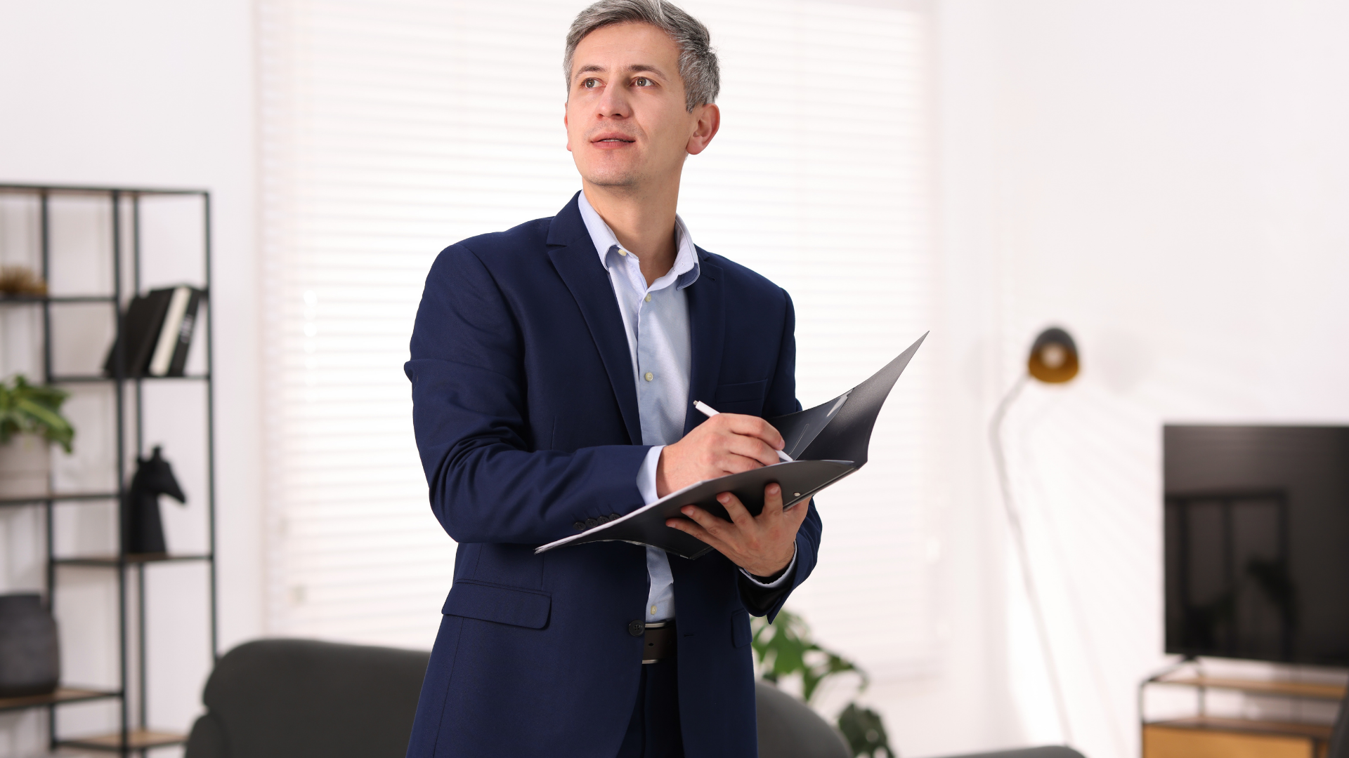 A person in a suit stands in a bright room, looking upward while writing in a black folder held in their hands.