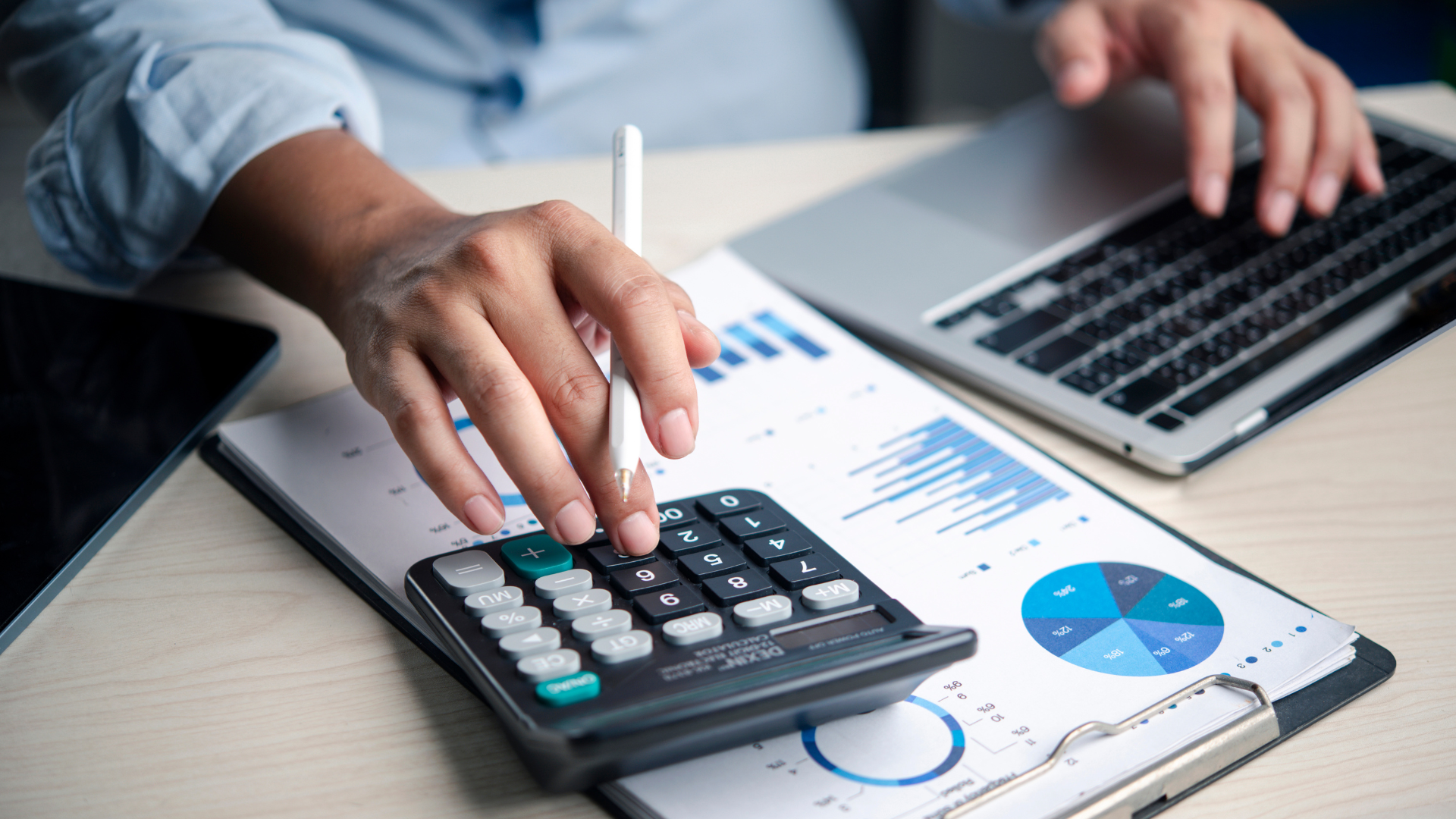 Person using a calculator and laptop, reviewing financial reports with charts on a desk.