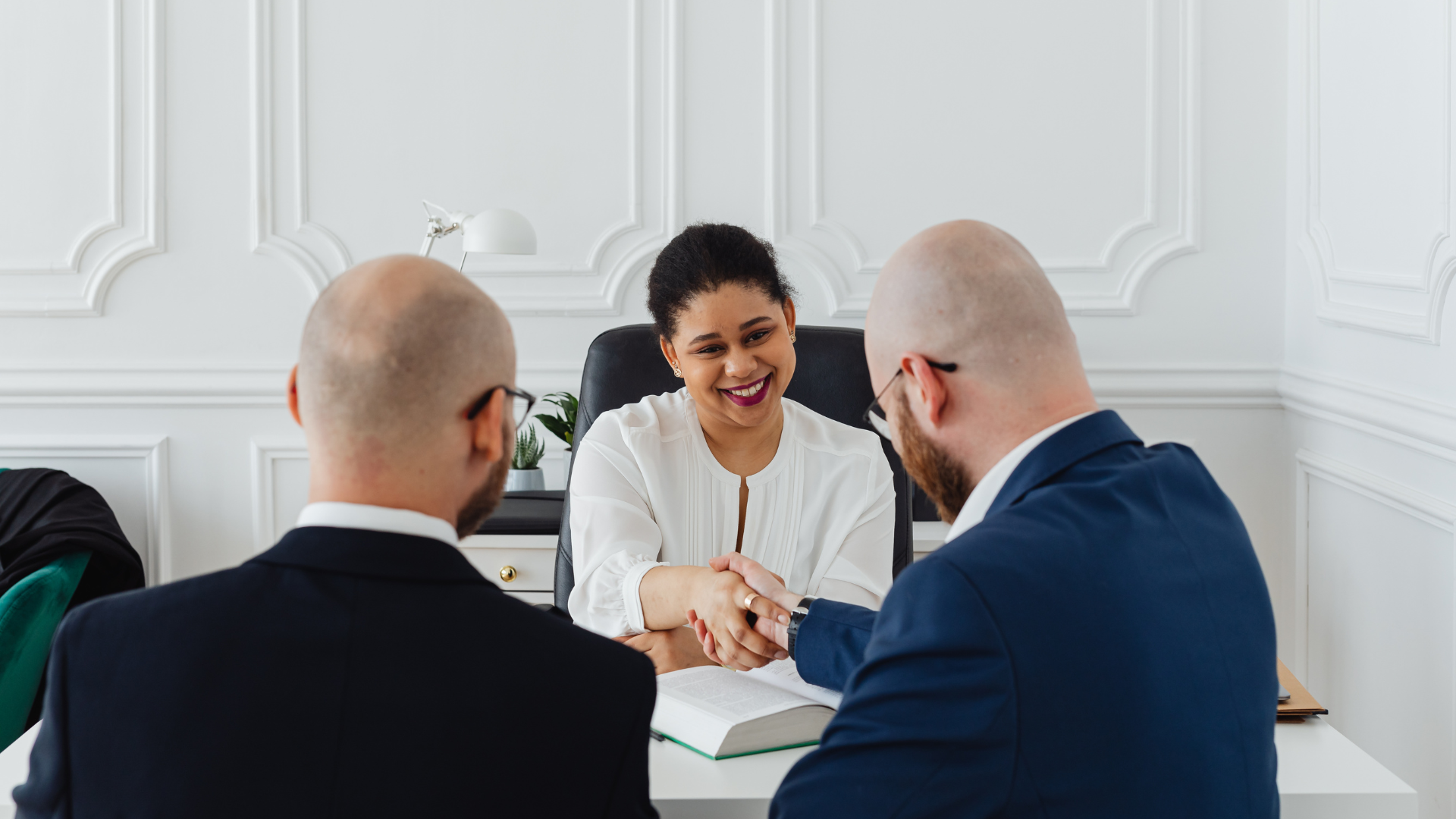 Two men in suits shaking hands with a smiling woman behind a desk in a white office.