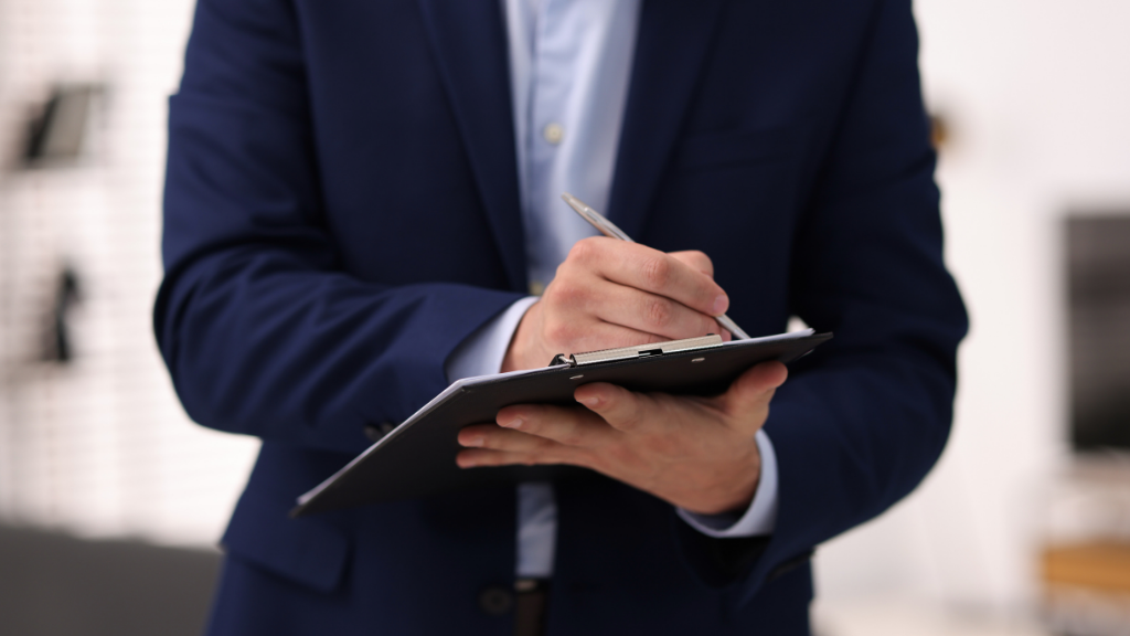 Person in a blue suit writing on a clipboard with a pen indoors.