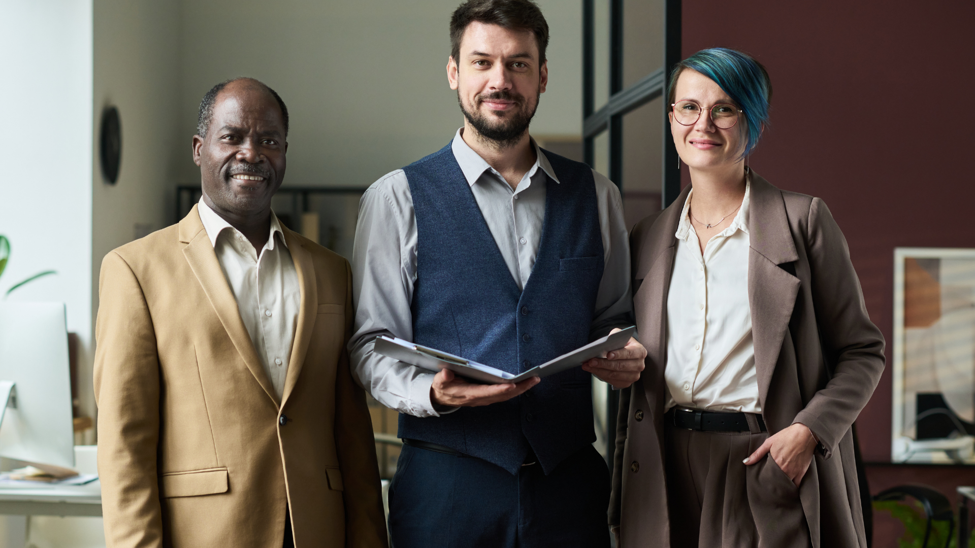 Three people posing together in an office. The man in the middle holds a binder, and they all smile.