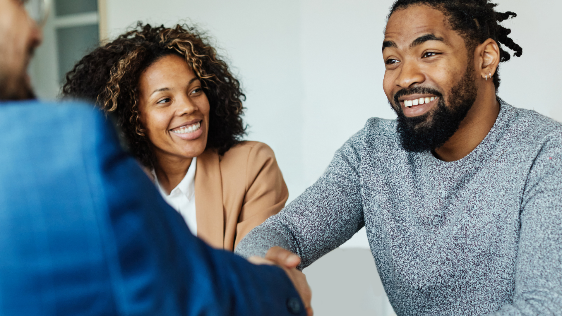 People shaking hands, smiling.  Man in blue suit, woman in tan blazer, and man in gray sweater.