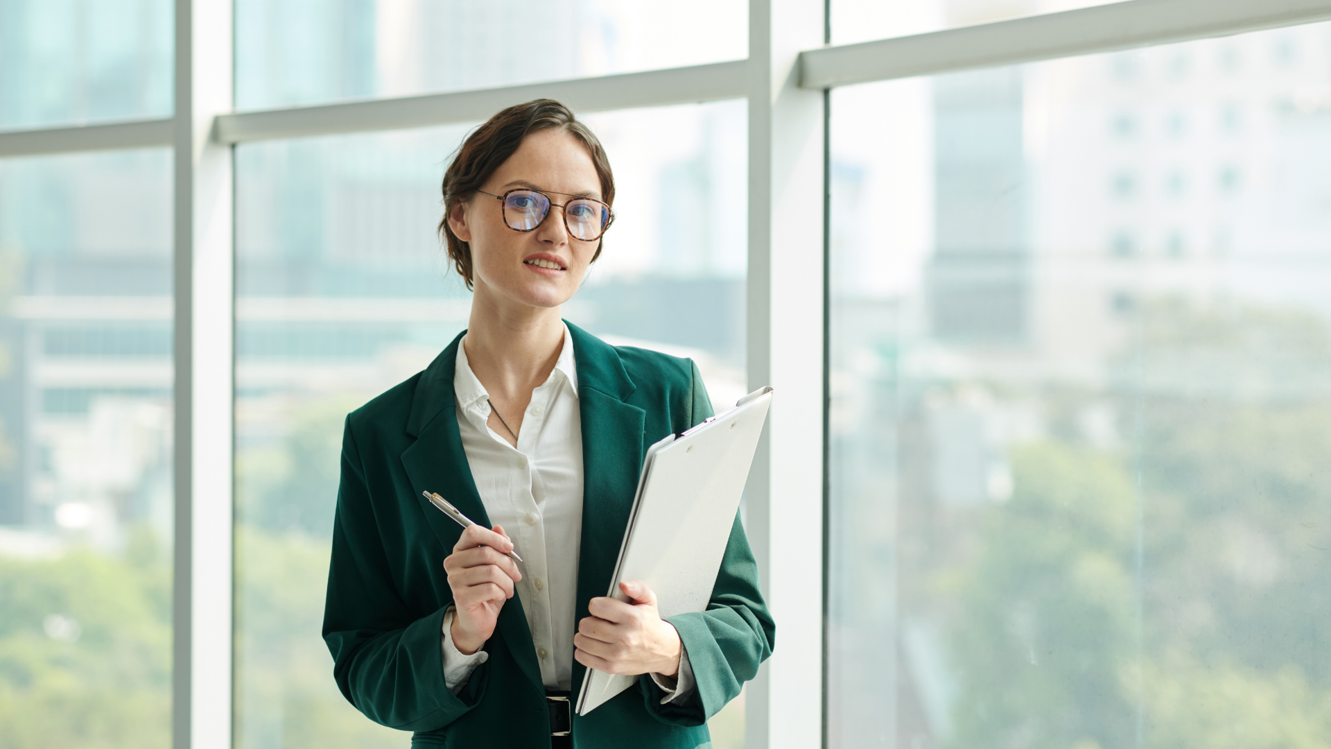 Woman in green blazer, glasses, and white shirt holding documents and pen by a window.