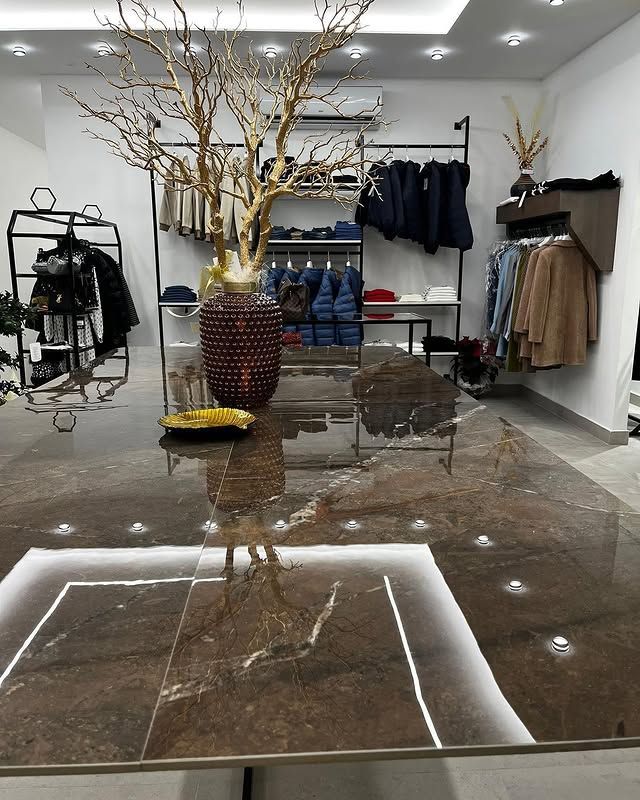 All'interno di un moderno e luminoso negozio di abbigliamento, un tavolo in marmo marrone lucido ospita un vaso scuro dalla texture particolare, ornato da rami dorati.