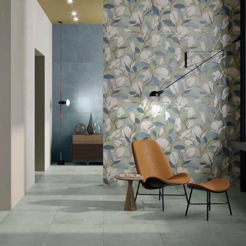 Una poltrona e un poggiapiedi sono posizionati su un pavimento di piastrelle grigie, addossati a una parete con carta da parati a motivi floreali, con un corridoio visibile sullo sfondo.