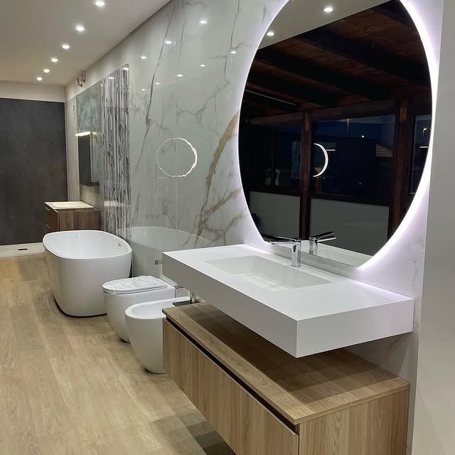 Un bagno moderno con mobile lavabo sospeso in legno con lavabo bianco, un grande specchio rotondo retroilluminato e una vasca da bagno indipendente.
