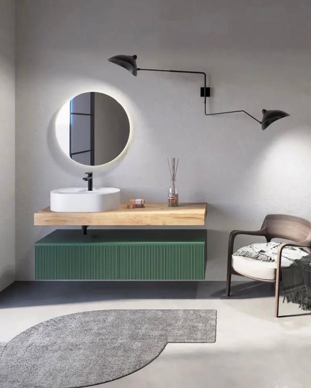 Mobile bagno moderno con ante a doghe verdi, ripiano in legno, lavabo bianco, specchio retroilluminato e lampada da parete accanto a una sedia.