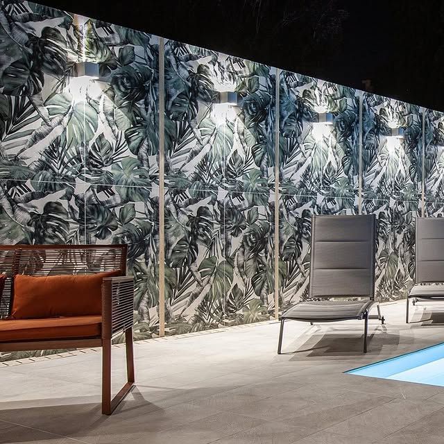 Un patio moderno di notte, con pannelli a parete decorati con motivi di foglie tropicali, una poltrona imbottita arancione e lettini grigi a bordo piscina.