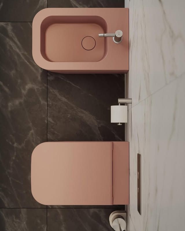 Vista dall'alto di un WC sospeso e di un bidet rosa opaco in un bagno con pareti rivestite in pietra scura e marmo bianco.
