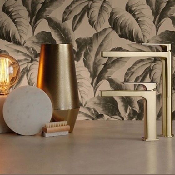 Due rubinetti color oro su un piano di lavoro, accompagnati da un vaso dorato, una pietra circolare bianca e una piccola spazzola.