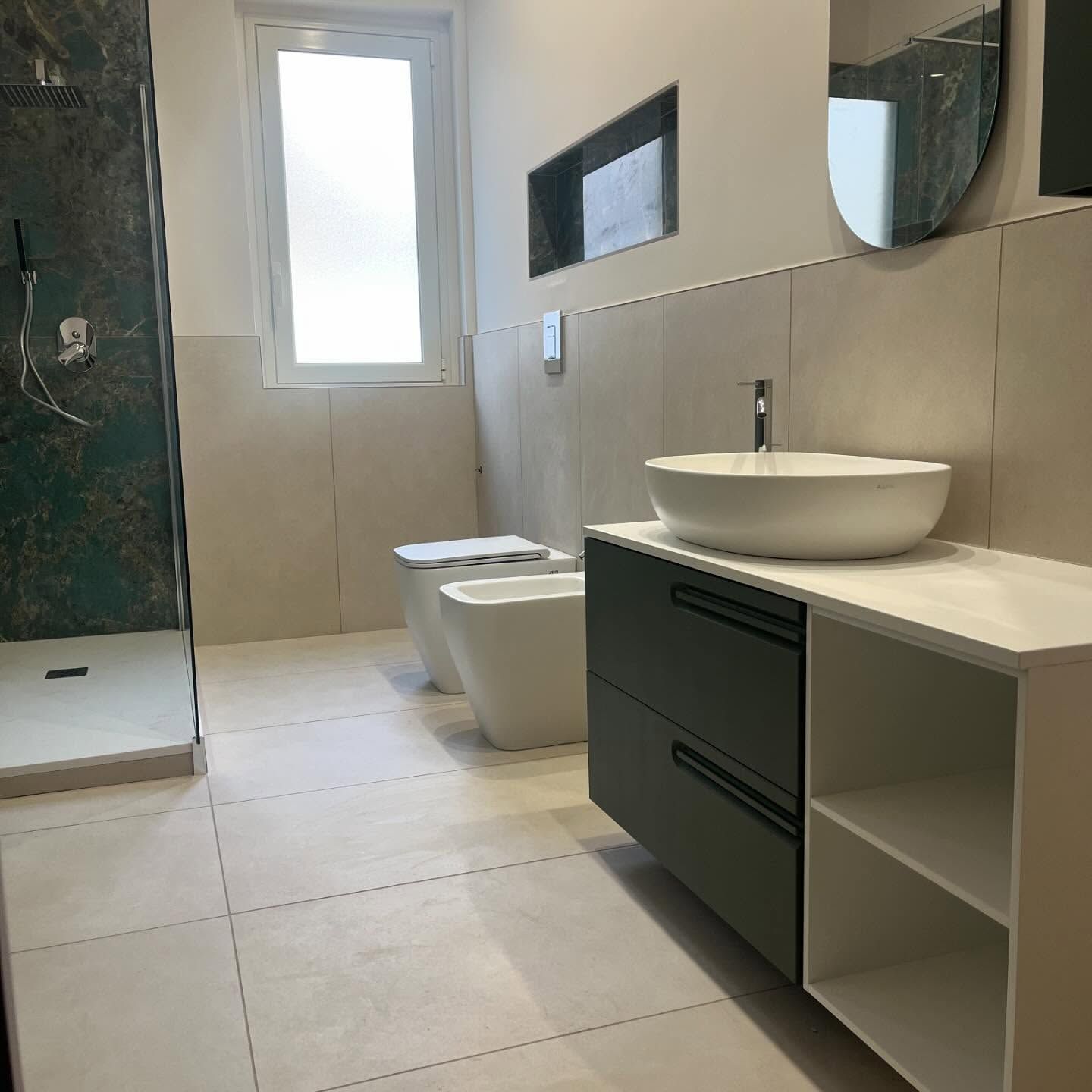 Un bagno moderno con lavabo a ciotola bianco su mobile scuro, bidet, WC e cabina doccia con pareti piastrellate.