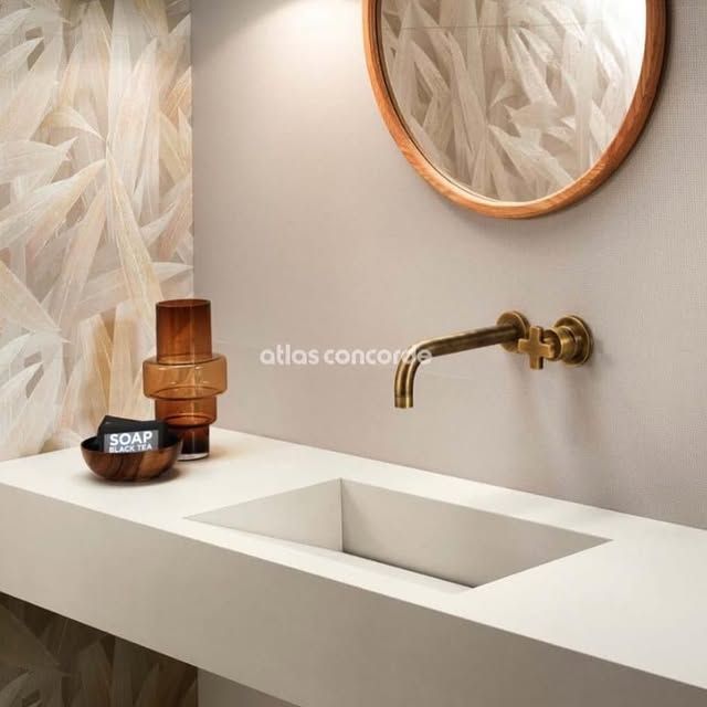 Un lavabo da bagno bianco e minimalista con rubinetto a parete in ottone, specchio con cornice in legno e dispenser di sapone in vetro ambrato.