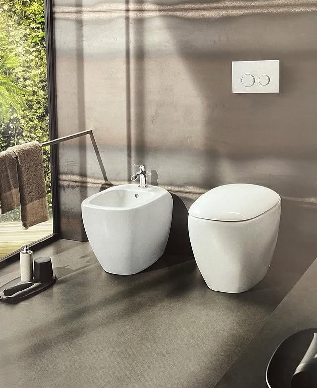 Set moderno composto da bidet bianco e WC sospeso in un bagno con pareti scure e texturizzate e un'ampia finestra.