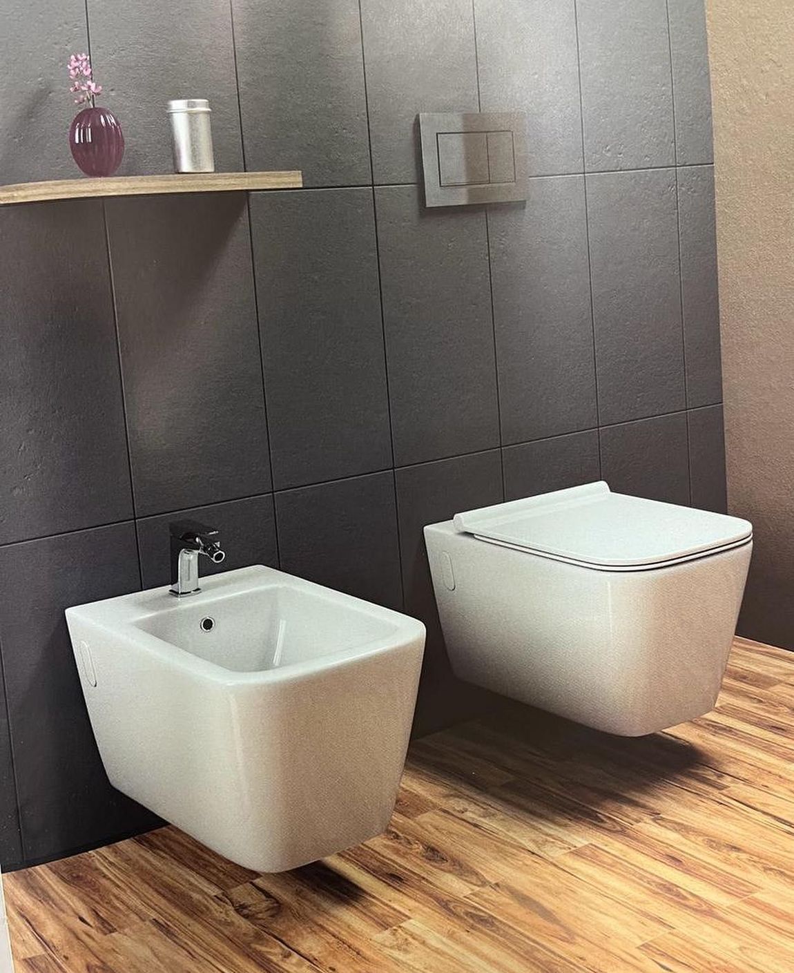 Bidet e WC bianchi a parete, incastonati in una parete rivestita di piastrelle grigio scuro sopra un pavimento effetto legno.