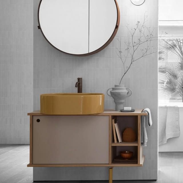 Mobile bagno moderno con lavabo a ciotola giallo, struttura in legno chiaro, mobiletto color tortora opaco e specchio rotondo a parete.