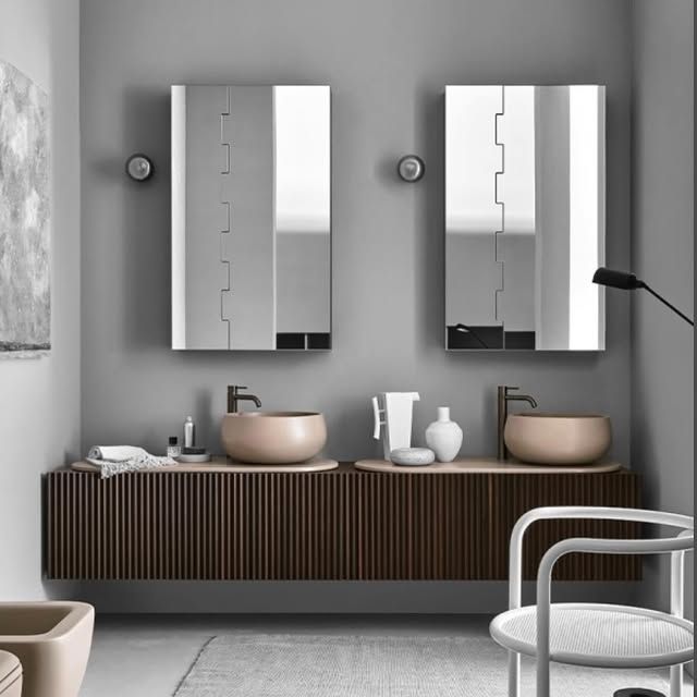 Un moderno doppio lavabo con lavandini circolari beige, mobili scuri scanalati e specchi rettangolari su una parete grigia.