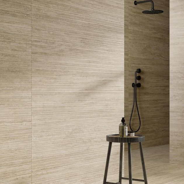 Un bagno moderno con pareti in pietra beige texturizzata, un miscelatore doccia scuro e uno sgabello minimalista in primo piano.