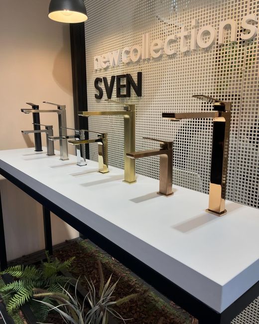 Una selezione di rubinetti da bagno moderni del marchio SVEN, disponibili in diverse finiture metalliche, esposti su un piano bianco di fronte a una parete piastrellata.