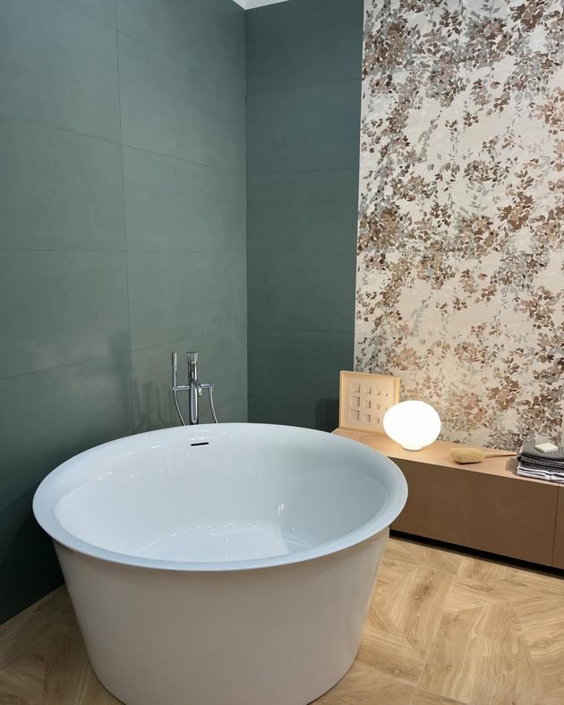In una stanza con pareti rivestite di piastrelle verdi, carta da parati a motivi floreali e pavimento in legno chiaro, si trova una vasca da bagno bianca e circolare autoportante.