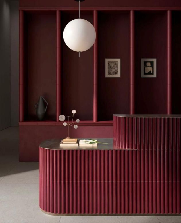 Un moderno bancone reception color bordeaux con un design scanalato si trova di fronte a scaffalature a muro in tinta, di un rosso intenso, e a una lampada a globo.