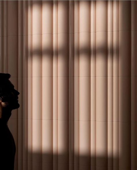 Sagoma di profilo di una persona che osserva la luce che filtra da una finestra, creando un'ombra a forma di croce su una parete scanalata.