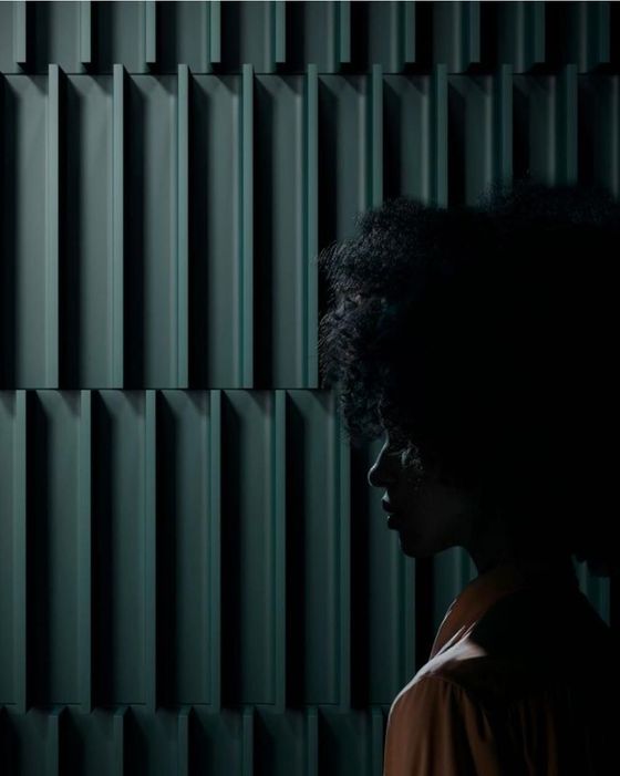 Una figura umana, in controluce, si staglia contro una parete scura a coste verticali, illuminata da una luce che ne mette in risalto il profilo.