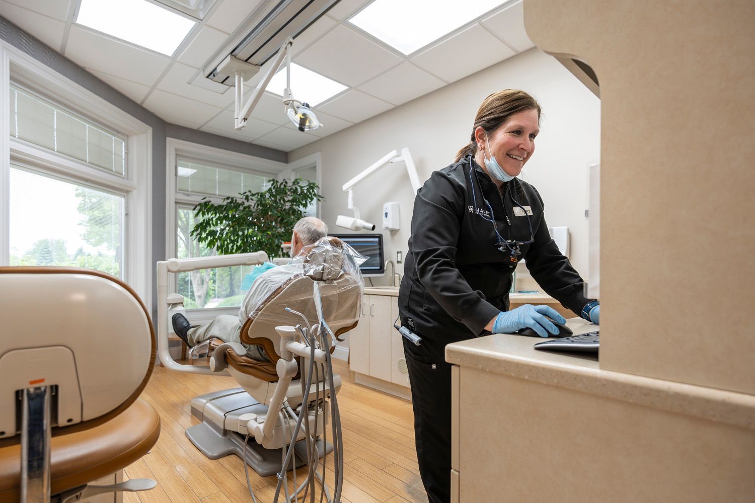 A dental hygienist is standing in a dental office next to a man in a dental chair at Maley Family Dental