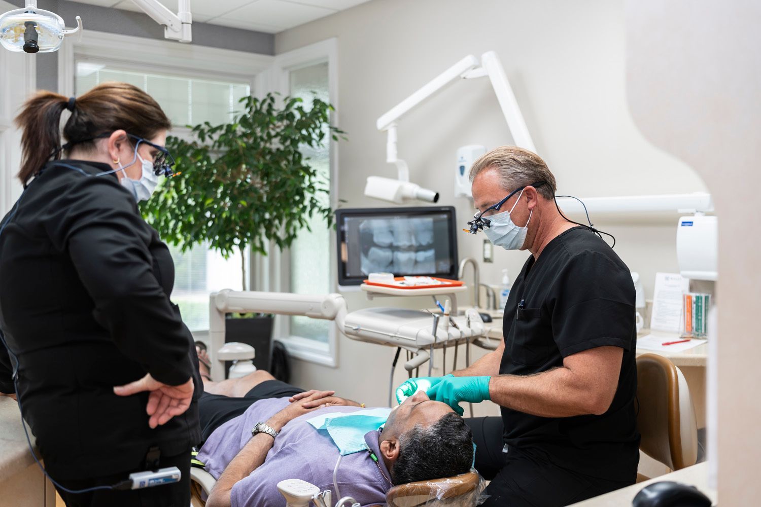 A man is laying in a dental chair while Dr. Maley and a dental hygienist examines his teeth at Maley Family Dental