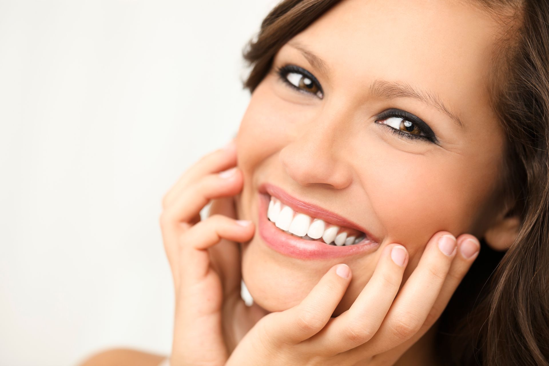 Woman smiling, holding her cheeks, with bright white teeth and dark eye makeup.