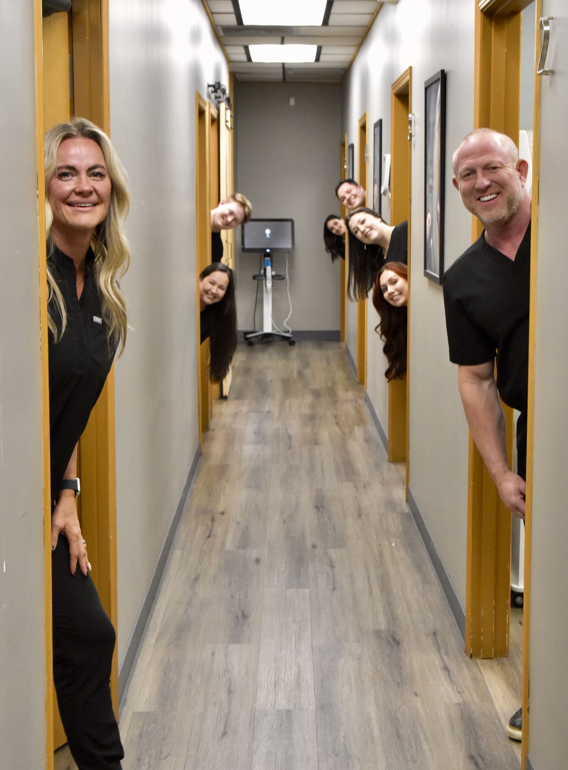 People in scrubs peeking from doorways down a hallway. One woman smiles directly at the camera.